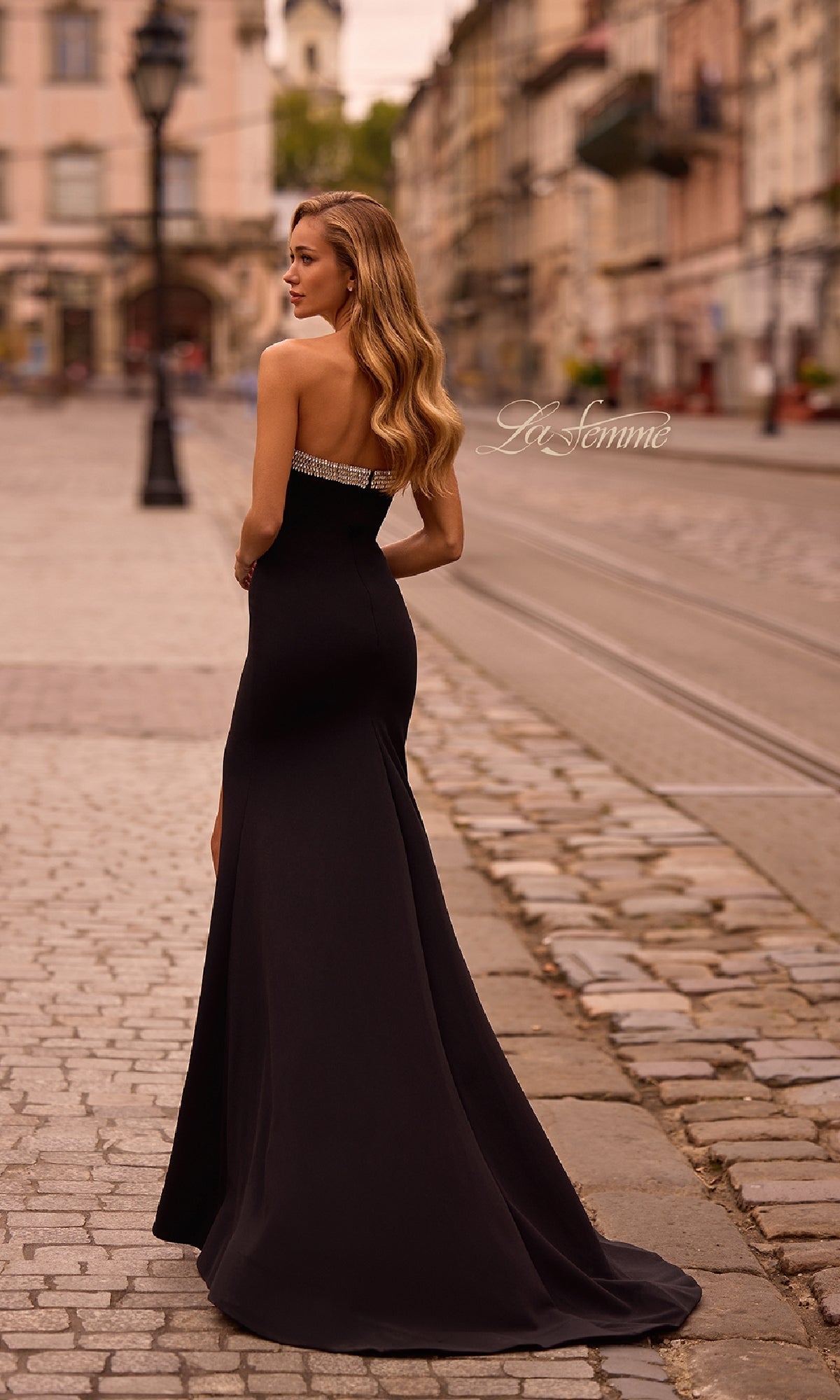 A woman with long hair wears the Bead-Trimmed Long Prom Dress: La Femme 32891, standing on a cobblestone street in the city.